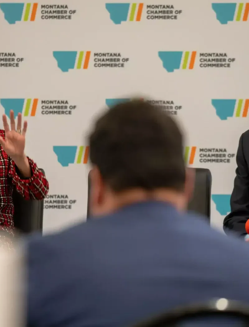 Secretary Christi Jacobsen discusses her 1st term successes and 2nd term outlook, speaking with a yellow microphone beside a man with a red microphone at a Montana Chamber of Commerce event, as an audience member listens in the foreground.