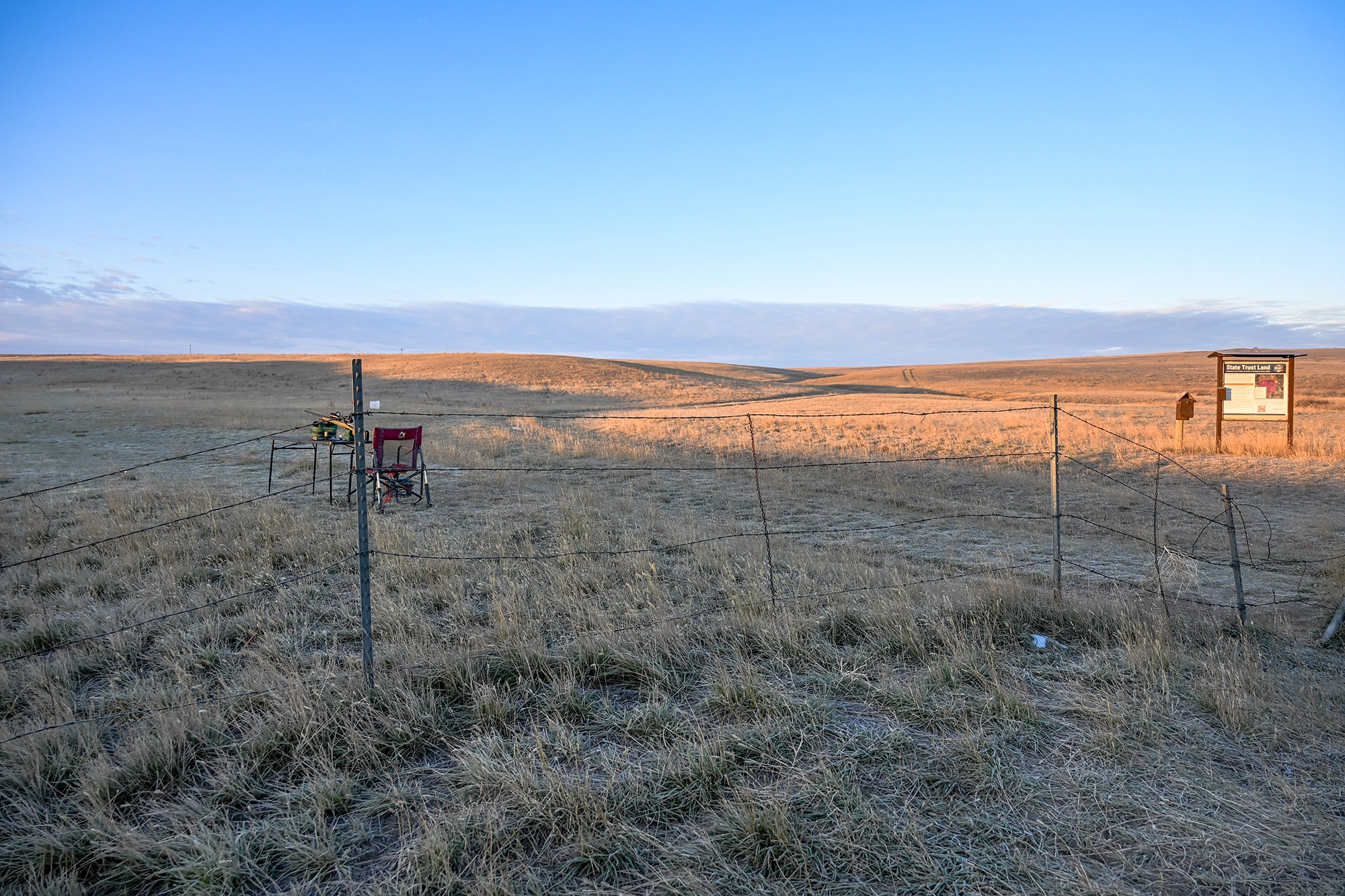 A folding chair and small table with a rifle sit behind a fence at the site of the proposed Logan Public Shooting Range in the Gallatin Valley, with a State Trust Land sign visible in the background.