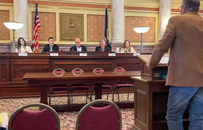 A man speaks at a podium to the Land Board panel of five officials in a formal room with ornate decor, American and state flags, and red chairs. The officials listen attentively as he addresses public access to the Big Snowy Mountains WMA.