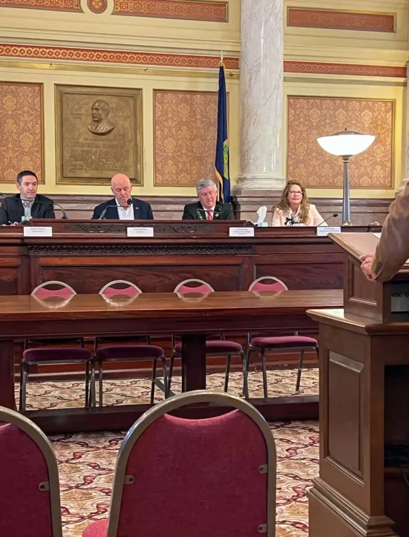 A man speaks at a podium to the Land Board panel of five officials in a formal room with ornate decor, American and state flags, and red chairs. The officials listen attentively as he addresses public access to the Big Snowy Mountains WMA.
