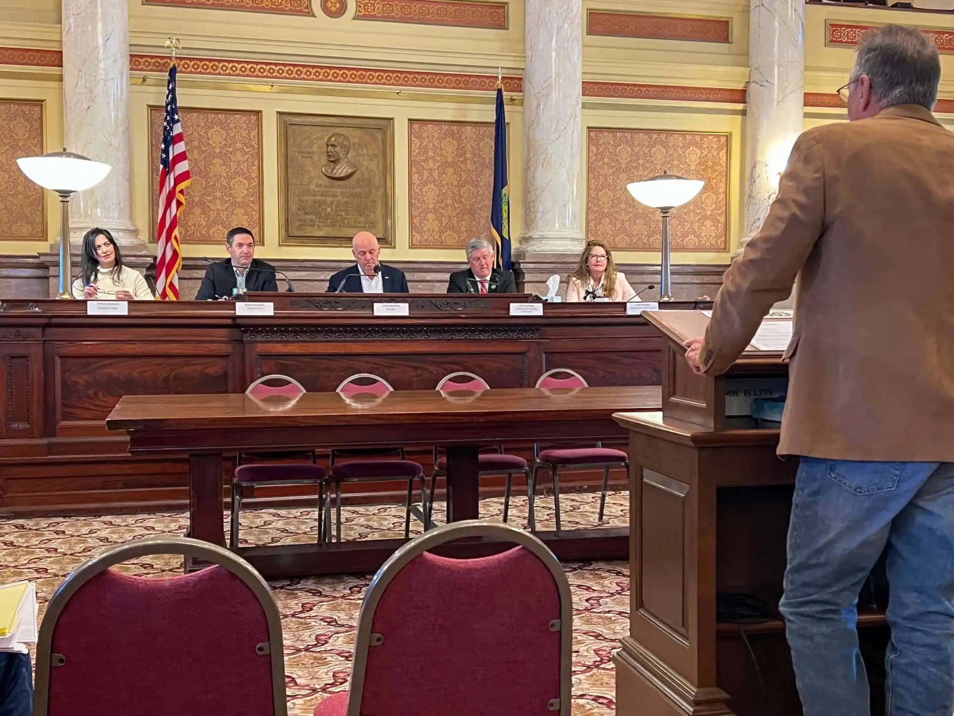 A man speaks at a podium to the Land Board panel of five officials in a formal room with ornate decor, American and state flags, and red chairs. The officials listen attentively as he addresses public access to the Big Snowy Mountains WMA.