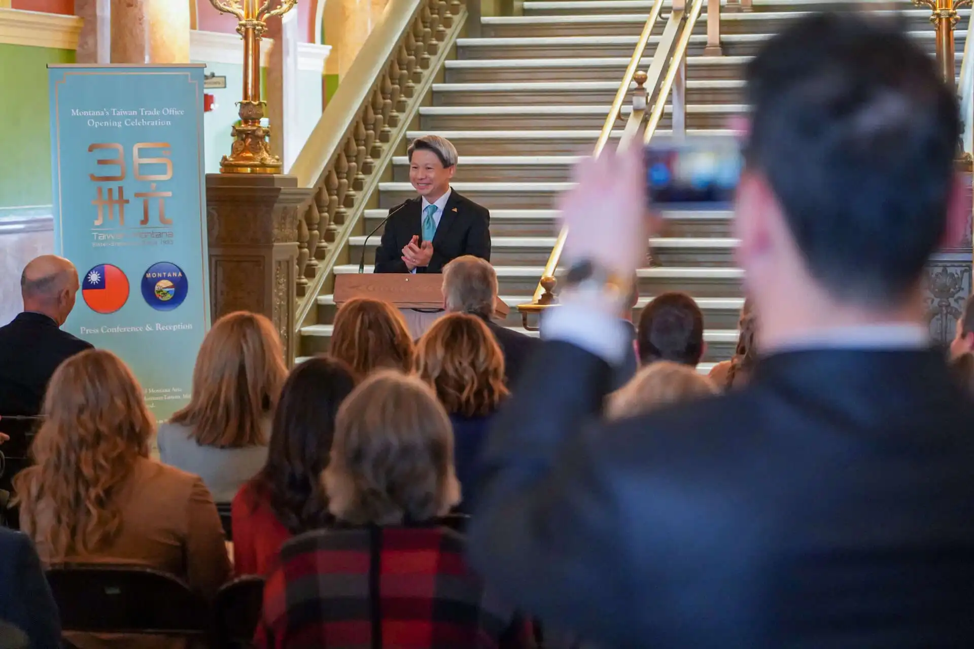 A person speaks at a podium to an audience in a formal setting, while another person in the foreground takes a photo or video with a phone. A banner with Taiwan and Missouri symbols is visible on the left.