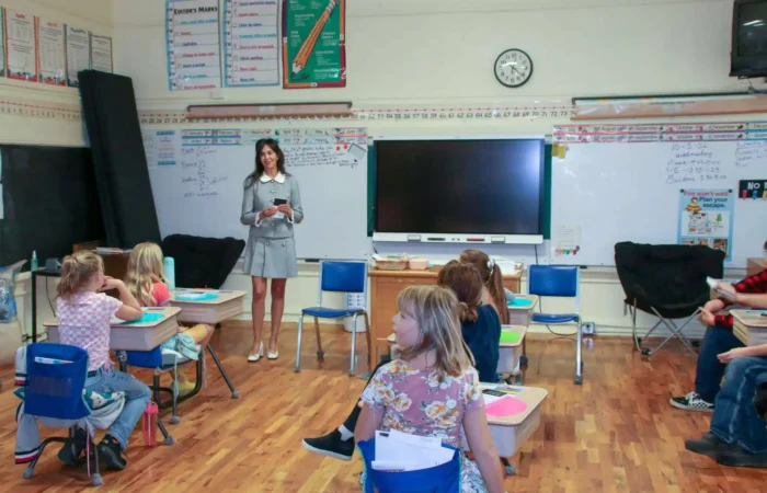 Secretary Christi Jacobsen discusses Constitutions with students in Twin Bridges, Willow Creek 8 A teacher stands at the front of a classroom, holding a book and smiling at a group of young students seated at desks. The classroom has a smartboard, posters, and writing on the whiteboard.