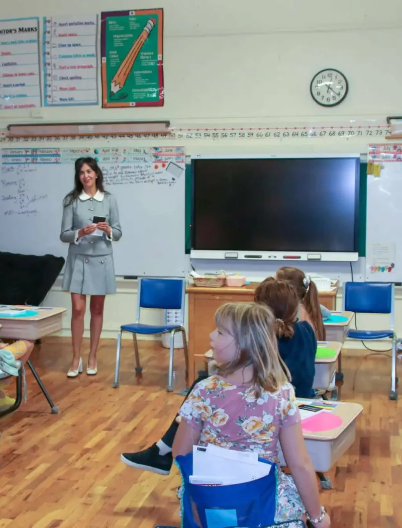 A teacher stands at the front of a classroom, holding a book and smiling at a group of young students seated at desks. The classroom has a smartboard, posters, and writing on the whiteboard.
