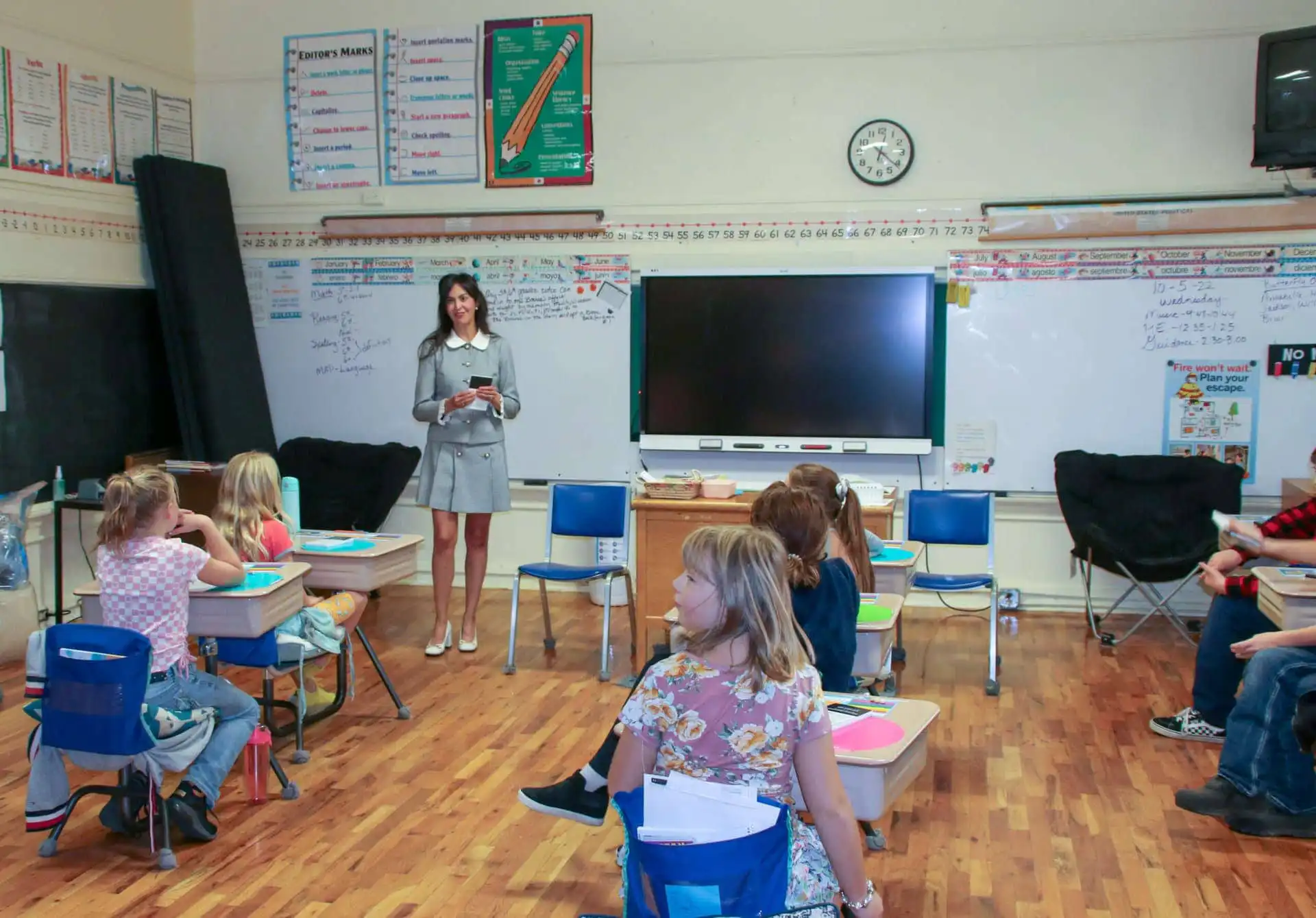 A teacher stands at the front of a classroom, holding a book and smiling at a group of young students seated at desks. The classroom has a smartboard, posters, and writing on the whiteboard.