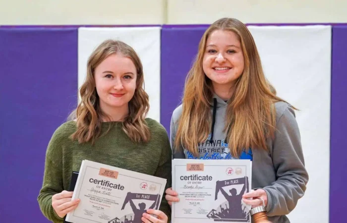 Winners announced in 2021 'What does the US Constitution mean to me' contest 4 Two young women, contest winners, stand side by side indoors, each holding a certificate and a trophy while smiling at the camera. The background features a purple and white wall.