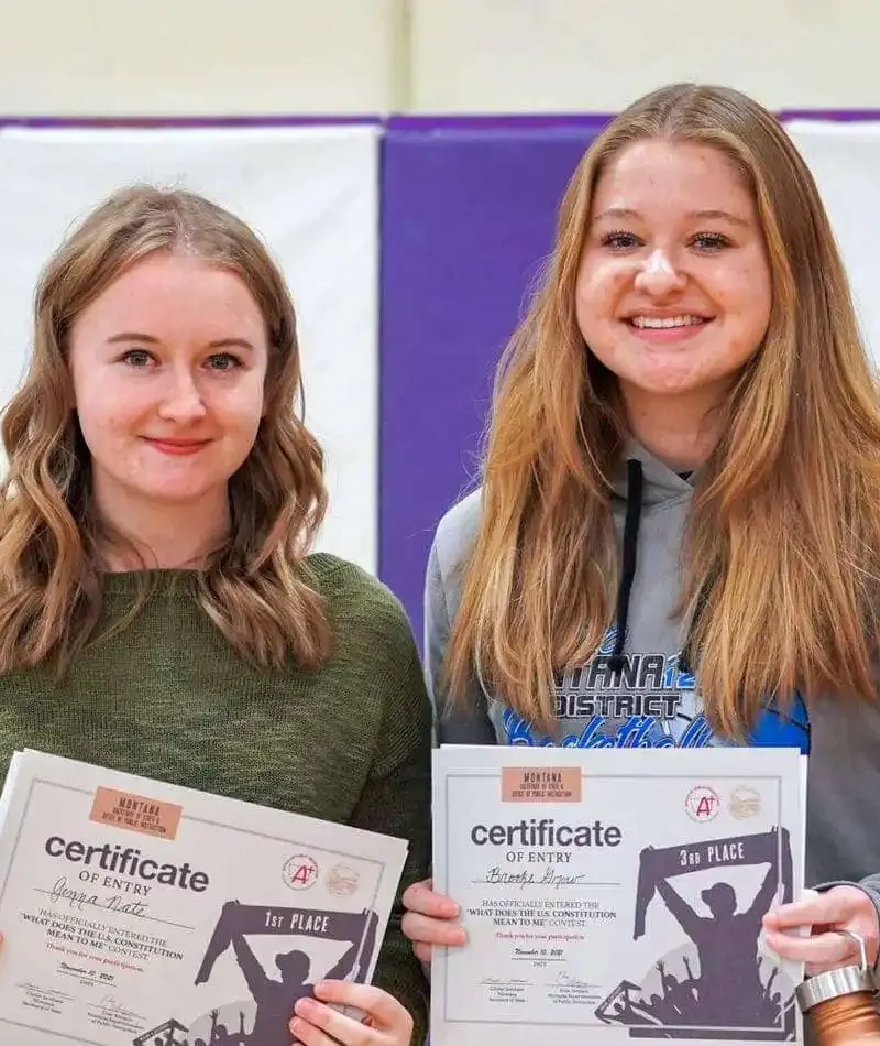 Two young women, contest winners, stand side by side indoors, each holding a certificate and a trophy while smiling at the camera. The background features a purple and white wall.