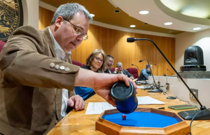 Montana State Board of Canvassers performs random-sample audit selection for 2024 General Election 2 A man in a brown blazer rolls a red die onto a blue tray at a meeting table during a random-sample audit, while others, including a woman in glasses, watch in the wood-paneled conference room with microphones.