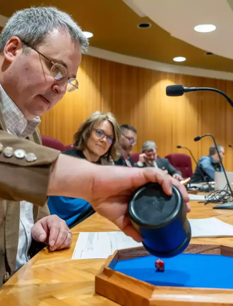 A man in a brown blazer rolls a red die onto a blue tray at a meeting table during a random-sample audit, while others, including a woman in glasses, watch in the wood-paneled conference room with microphones.