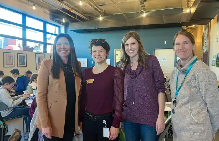 Secretary of State’s Business Services Division participates in Helena Public Schools PEAK Program 2 Four women are standing and smiling inside a brightly lit room with large windows at Helena Public Schools; people are working at tables in the background, highlighting the collaborative spirit of the PEAK Program.