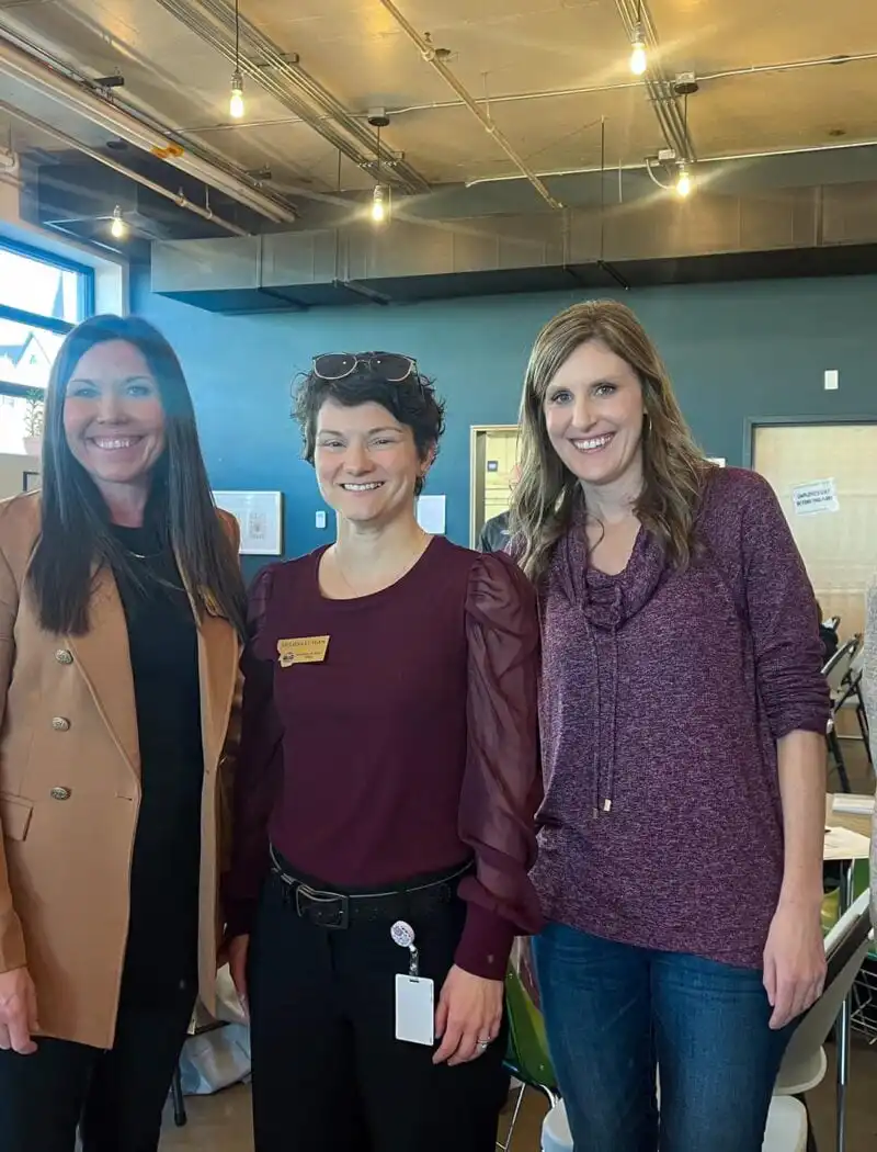 Four women are standing and smiling inside a brightly lit room with large windows at Helena Public Schools; people are working at tables in the background, highlighting the collaborative spirit of the PEAK Program.