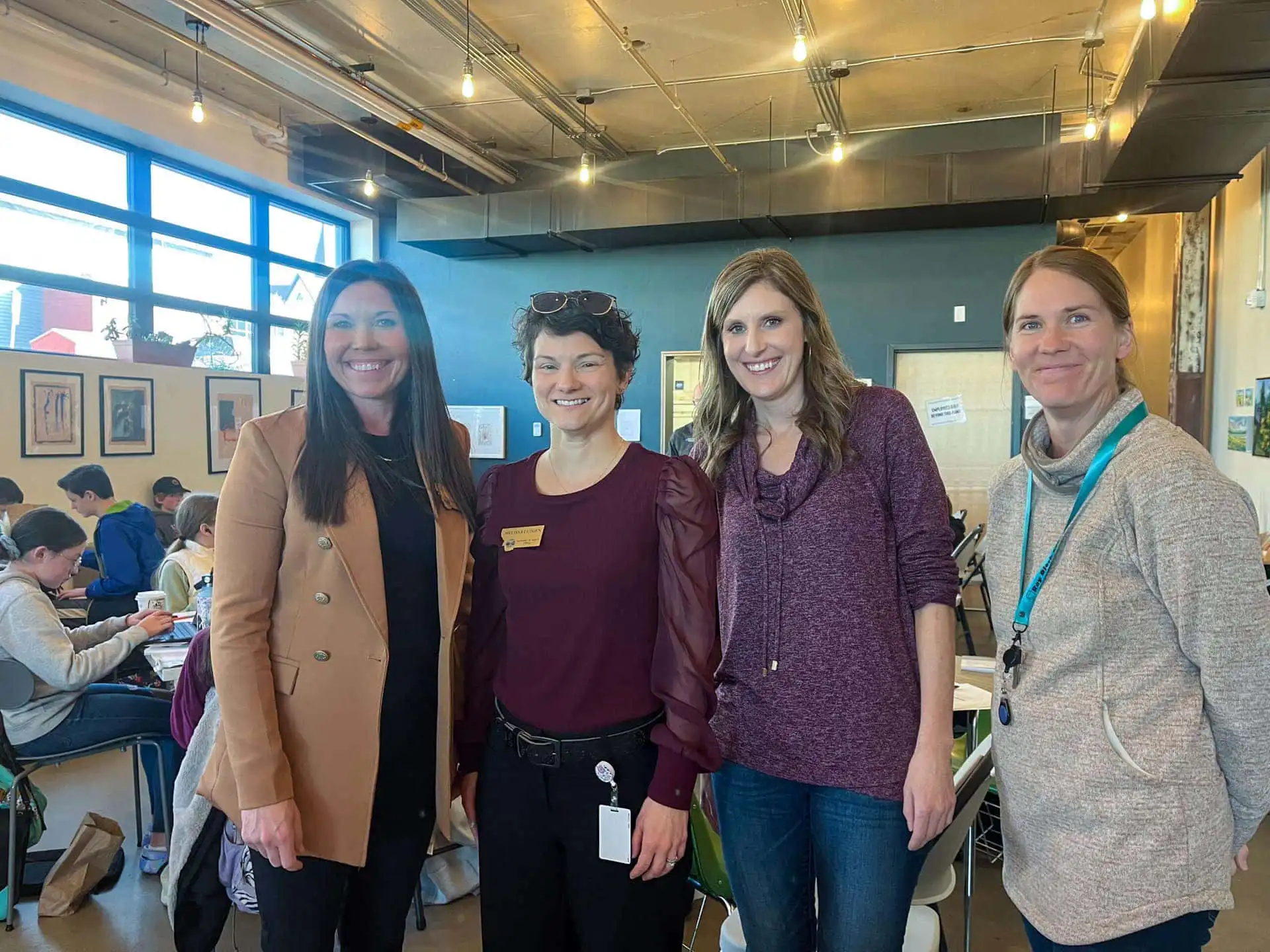 Four women smile in a brightly lit room with large windows and art on the walls. They represent the Business Services Division of the Secretary of State, gathering for a PEAK Program event amid modern decor and people seated at tables in the background.