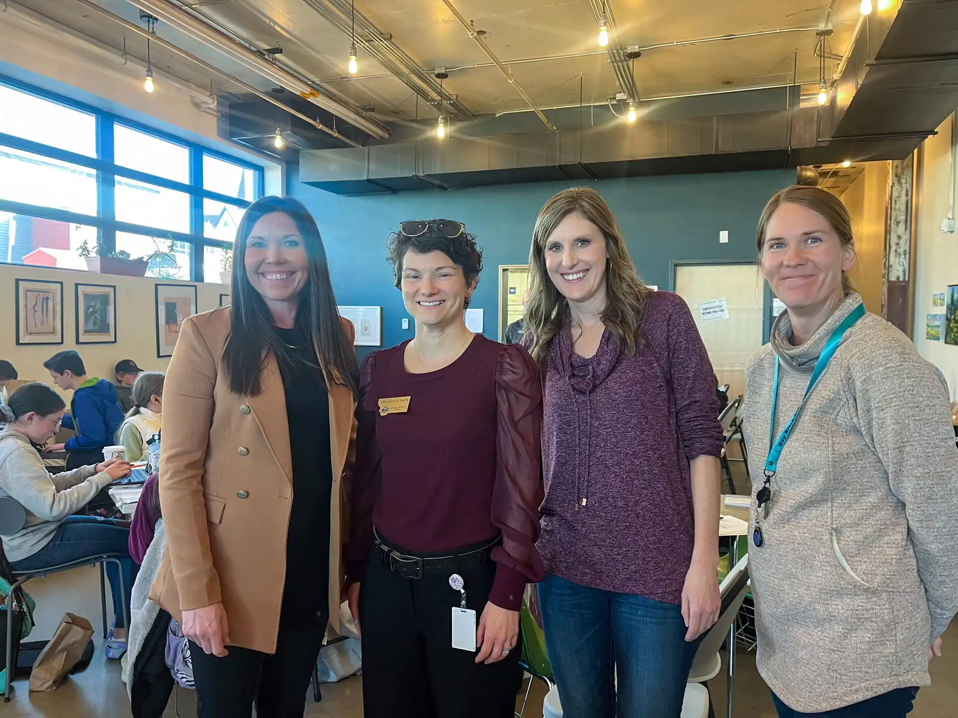 Four women are standing and smiling inside a brightly lit room with large windows at Helena Public Schools; people are working at tables in the background, highlighting the collaborative spirit of the PEAK Program.