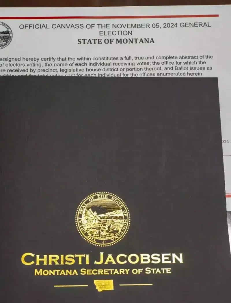 A black folder with a gold Montana state seal and Christi Jacobsen, Montana Secretary of State rests on top of an official state document for the November 5, 2024 general election.