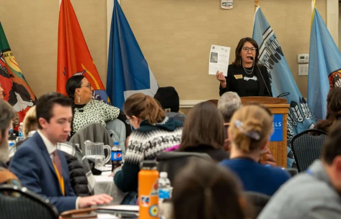 Secretary of State's Office staff attends Tribal Relations Training, thanks Governor's Office for hosting 4 A woman speaks at a podium, holding up a paper in front of an audience. People are seated at round tables, and various flags are displayed in the background.
