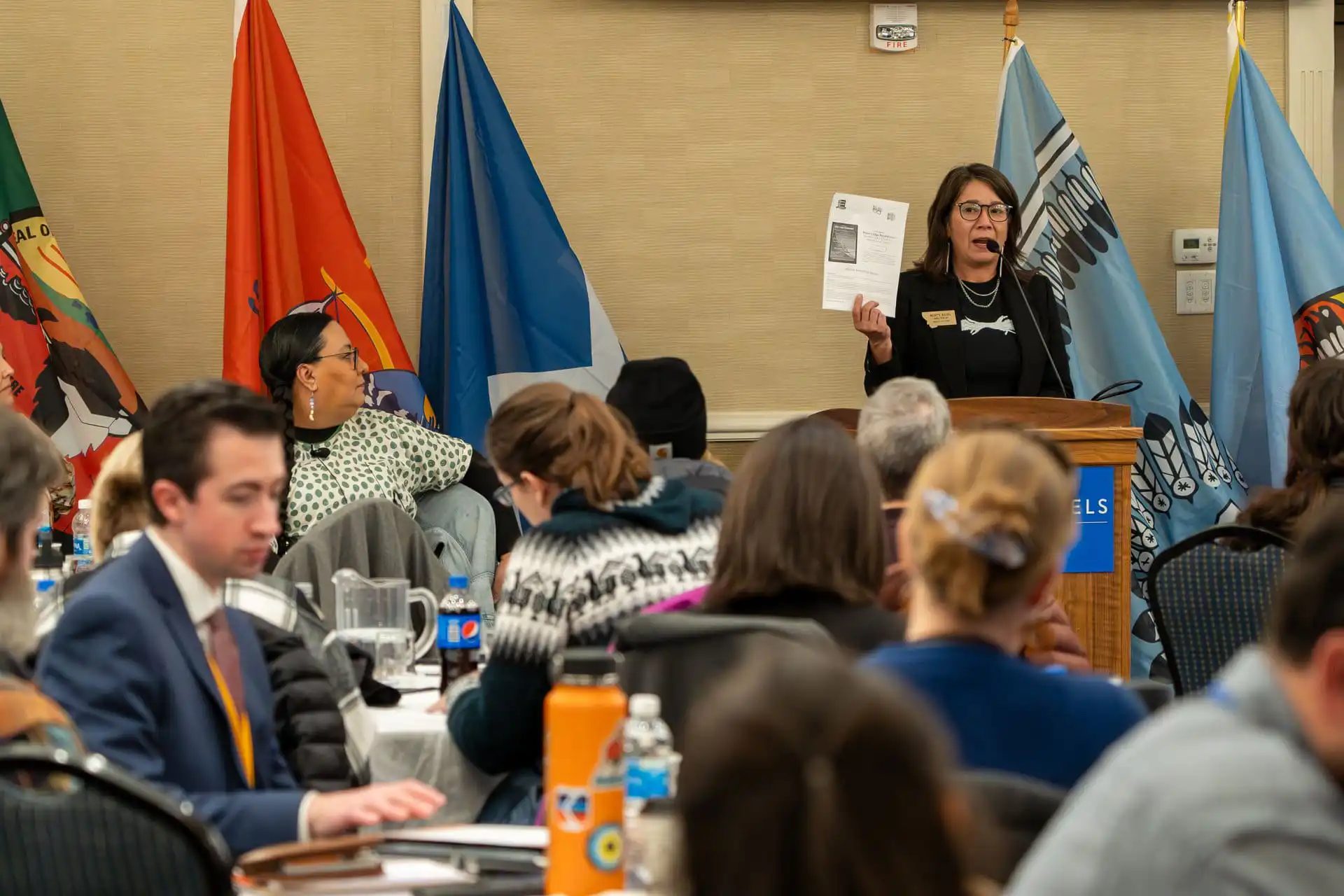 A woman stands at a podium holding up a paper and speaking to a seated audience at a Tribal Relations Training conference. Colorful flags are displayed behind her, and attendees are listening and taking notes.