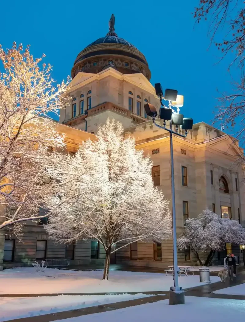 A historic building with a domed roof, illuminated at dusk and surrounded by snow-covered trees, stands serene. The calm winter evening is reminiscent of new businesses registered by Secretary Christi Jacobsen, marking growth amid frosted tranquility.