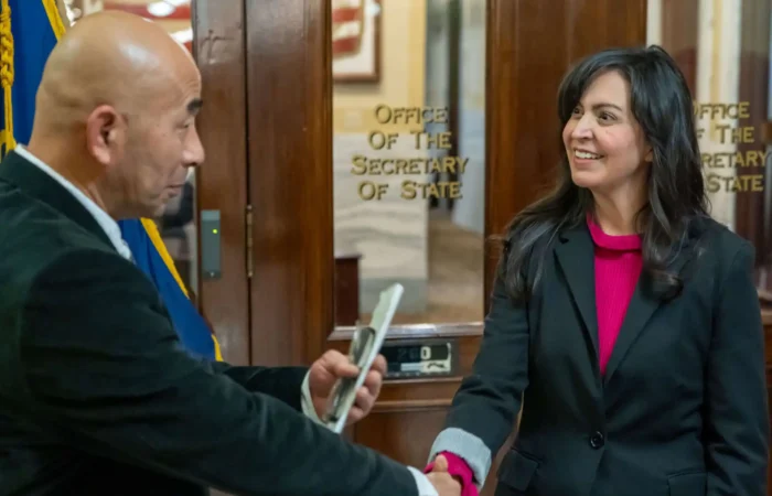 Secretary Christi Jacobsen meets with diplomatic delegation from Kumamoto Prefecture 8 A man and a woman, part of a diplomatic delegation, shake hands and smile in front of a glass door labeled Office of the Secretary of State. The man holds a phone, while the woman with long dark hair wears a blazer.