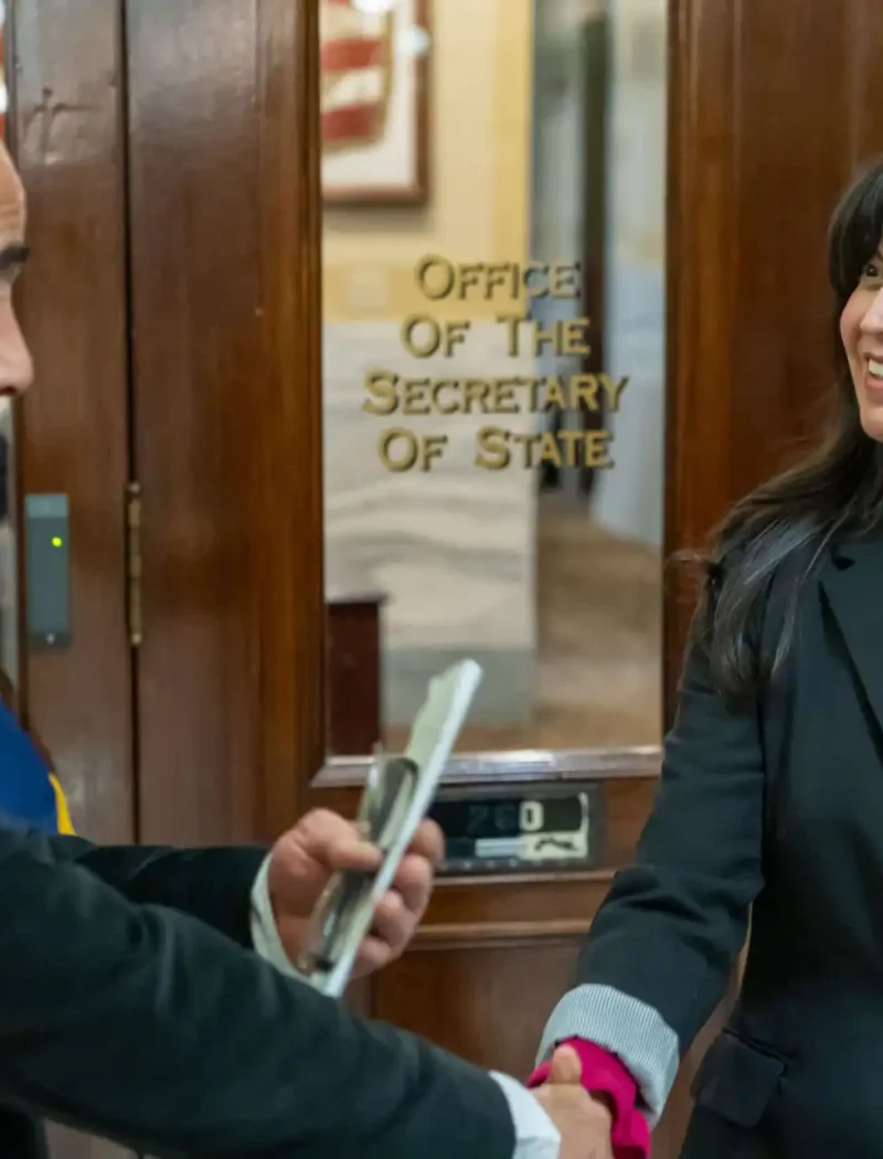 A man and a woman, part of a diplomatic delegation, shake hands and smile in front of a glass door labeled Office of the Secretary of State. The man holds a phone, while the woman with long dark hair wears a blazer.
