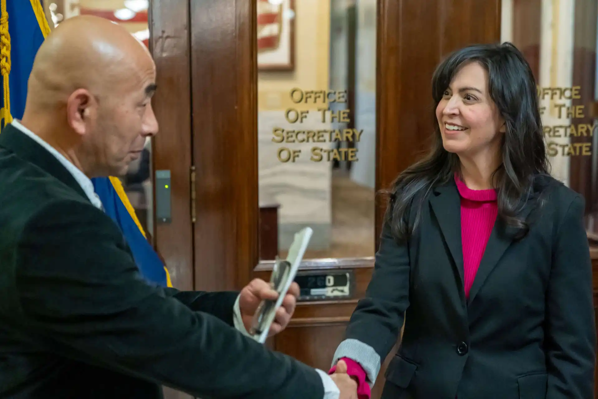 Secretary Christi Jacobsen meets with diplomatic delegation from Kumamoto Prefecture 6 A man and a woman, part of a diplomatic delegation, shake hands and smile in front of a glass door labeled Office of the Secretary of State. The man holds a phone, while the woman with long dark hair wears a blazer.