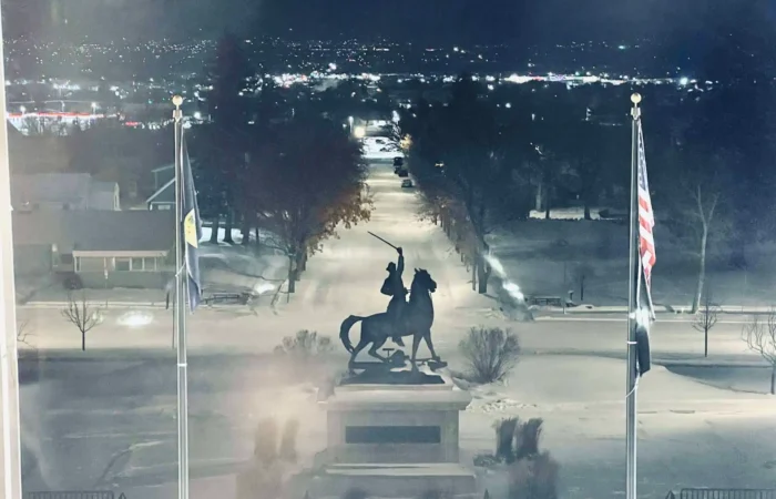 State Board of Canvassers conducts random-sample audit selection 8 A nighttime scene shows a statue of a rider on horseback holding a raised sword, flanked by two flagpoles with flags, on snowy ground where city lights twinkle in the distance—much like the careful audit selection by the State Board of Canvassers.