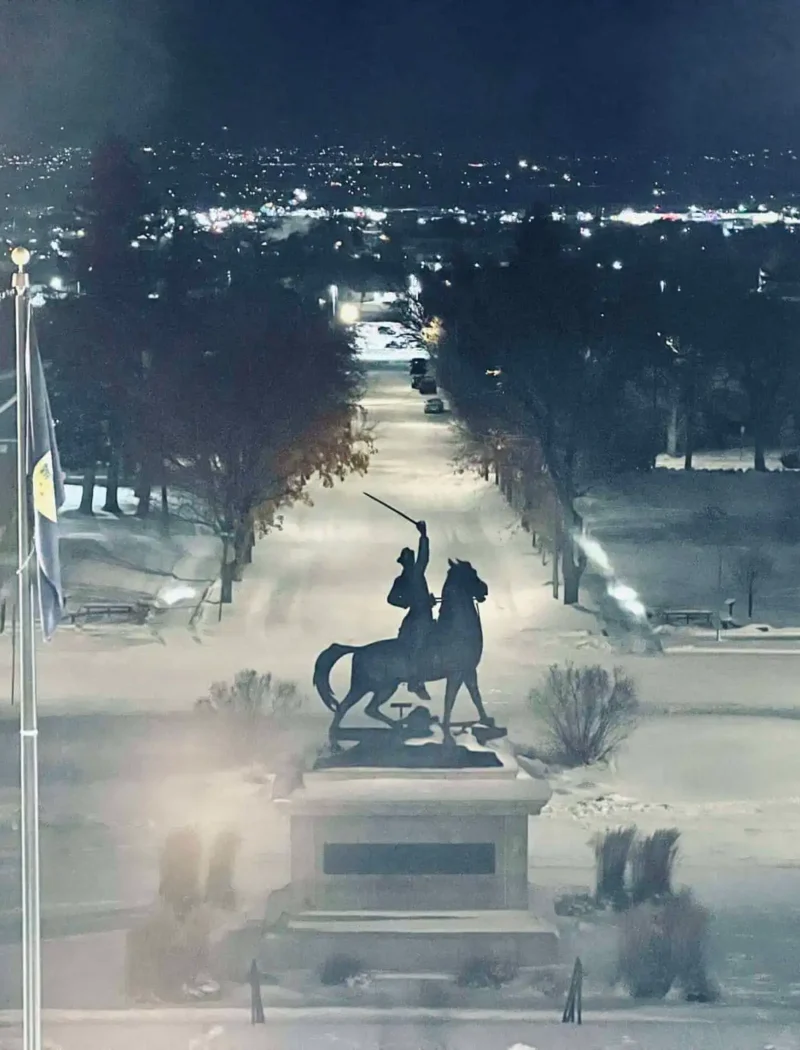 A nighttime scene shows a statue of a rider on horseback holding a raised sword, flanked by two flagpoles with flags, on snowy ground where city lights twinkle in the distance—much like the careful audit selection by the State Board of Canvassers.