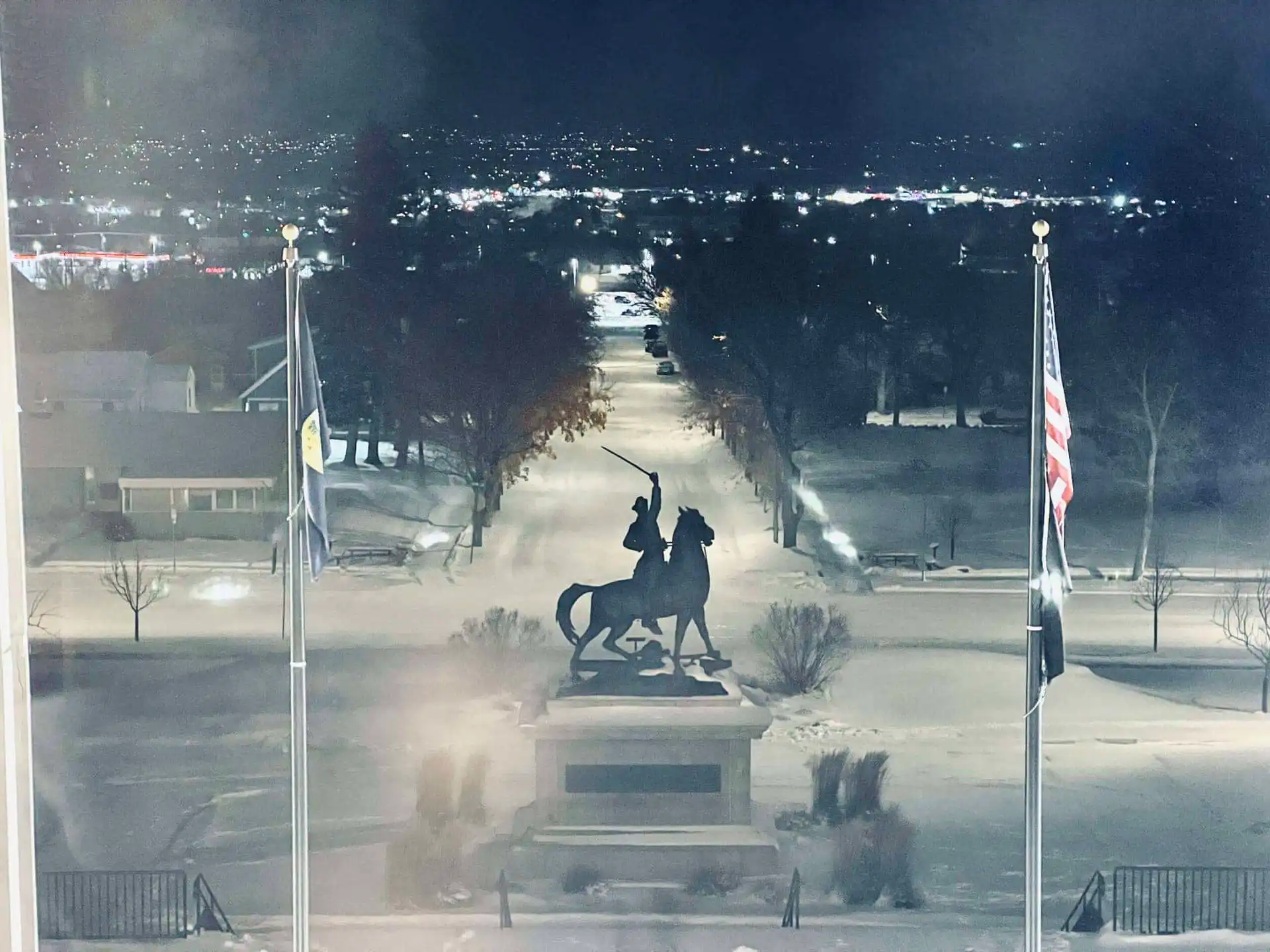A nighttime scene shows a statue of a rider on horseback holding a raised sword, flanked by two flagpoles with flags, on snowy ground where city lights twinkle in the distance—much like the careful audit selection by the State Board of Canvassers.