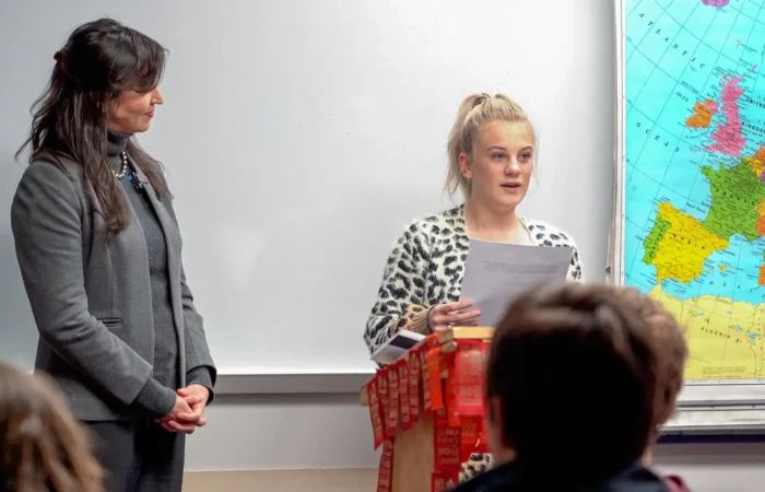 Belt High School senior collects 2nd-place in US Constitution Contest 10 A Belt High School senior reads from a sheet of paper at a podium decorated with red ribbons during the US Constitution Contest, while an older woman stands beside her. An audience watches, and a large map of Europe is visible on the wall behind them.