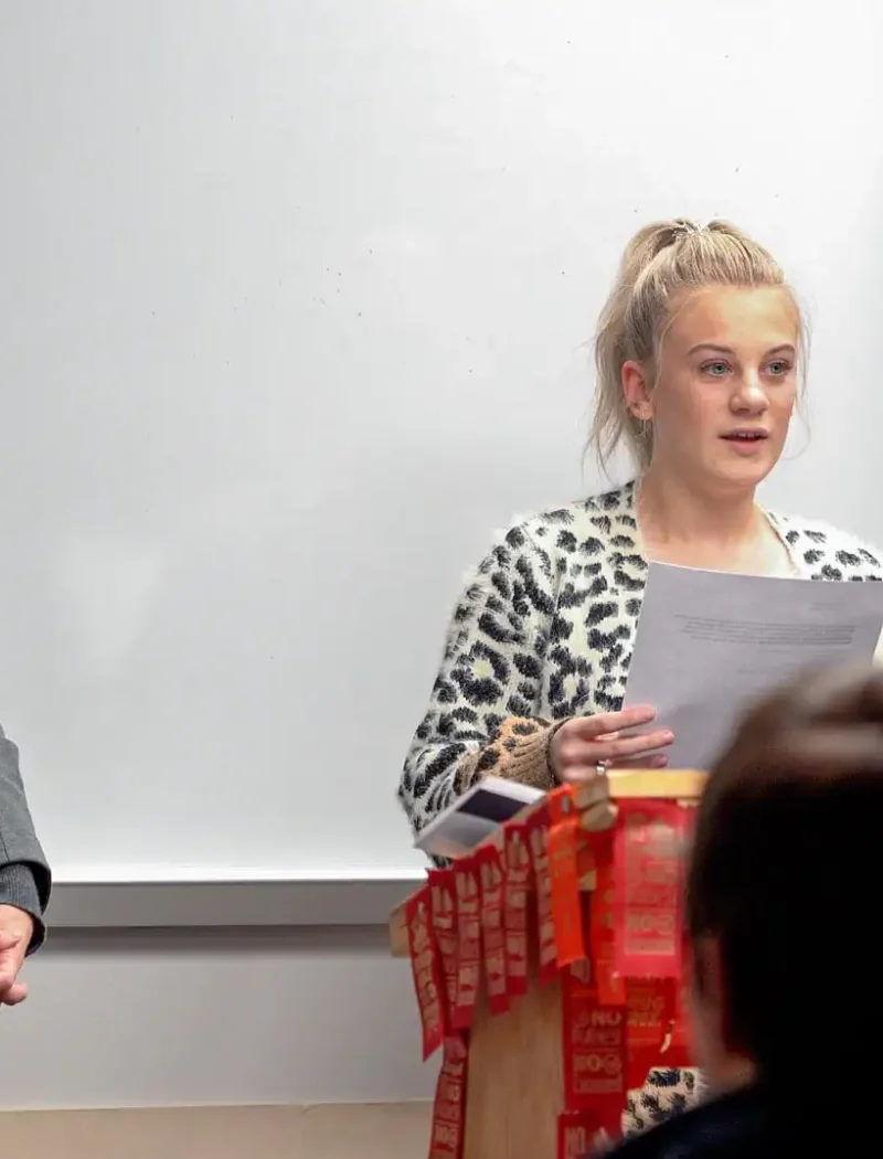 A Belt High School senior reads from a sheet of paper at a podium decorated with red ribbons during the US Constitution Contest, while an older woman stands beside her. An audience watches, and a large map of Europe is visible on the wall behind them.