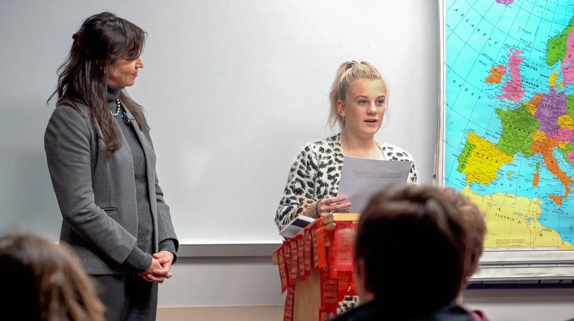 Belt High School senior collects 2nd-place in US Constitution Contest 10 A Belt High School senior reads from a sheet of paper at a podium decorated with red ribbons during the US Constitution Contest, while an older woman stands beside her. An audience watches, and a large map of Europe is visible on the wall behind them.