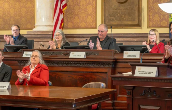 Secretary Christi Jacobsen: 'Montana's Electoral College votes cast for President Donald Trump' 10 A woman stands at a podium speaking while five people seated behind her and two at a table in front applaud. An American flag is displayed in the background of Montanas Electoral College meeting in a formal, wood-paneled room.