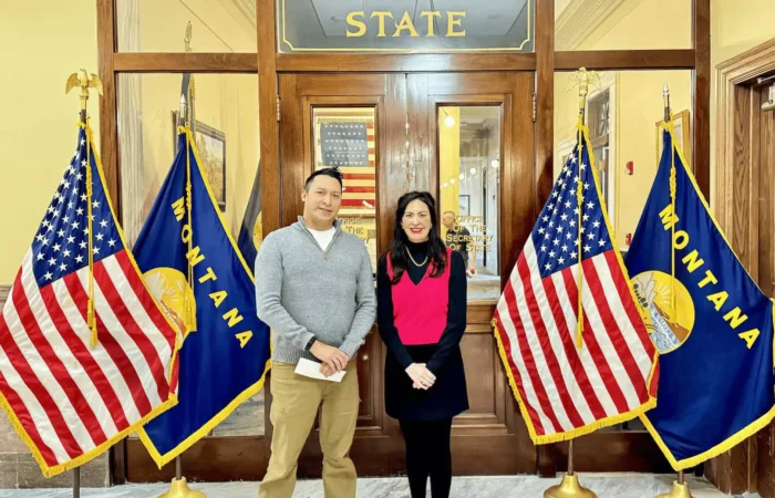 Secretary Christi Jacobsen: 'More than 240 candidate filings processed' 9 Two people stand smiling in front of a glass door labeled Secretary of State, where Secretary Christi Jacobsen has processed candidate filings, flanked by American and Montana flags in a formal building with marble floors.