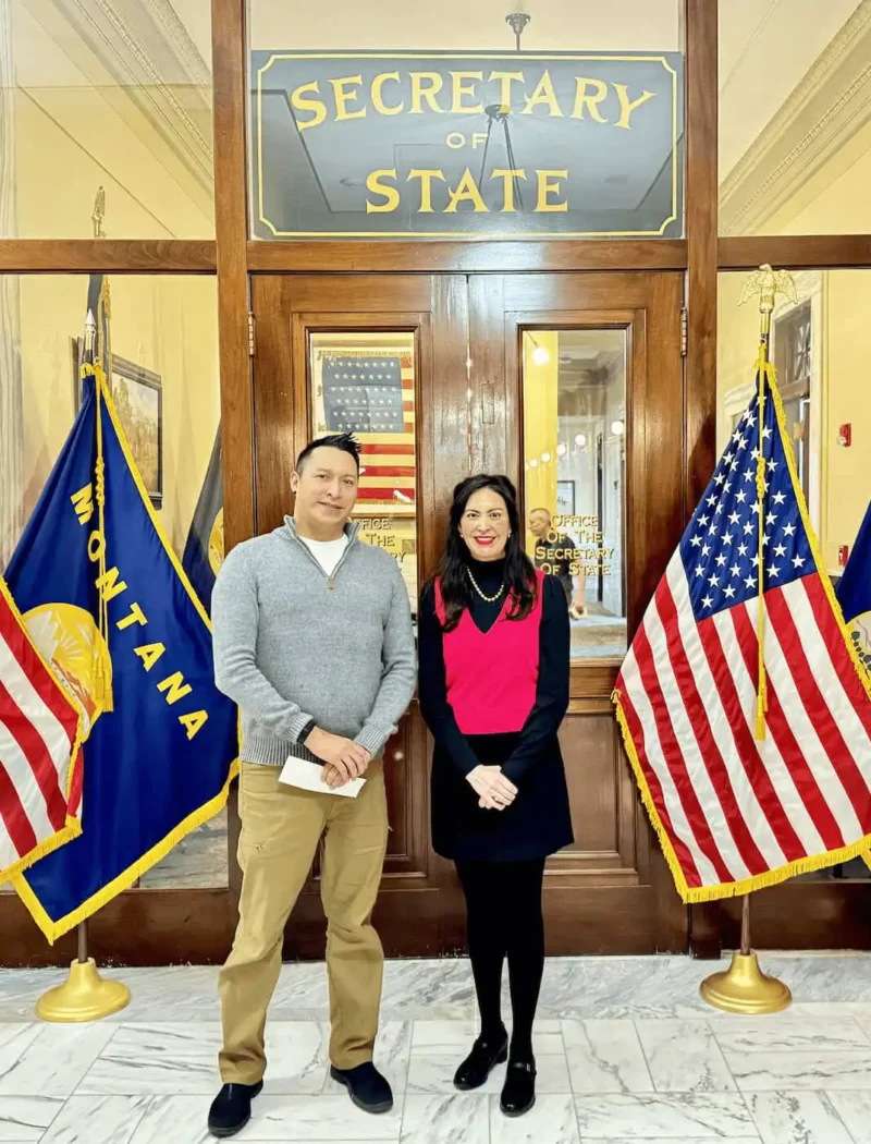 Two people stand smiling in front of a glass door labeled Secretary of State, where Secretary Christi Jacobsen has processed candidate filings, flanked by American and Montana flags in a formal building with marble floors.