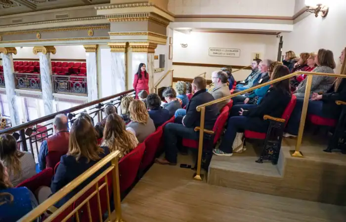 Secretary Christi Jacobsen: 'One month left to file 2025 annual report; fees waived for all Montana businesses' 10 A woman stands in front of a seated audience in a balcony area of an ornate building, possibly a theater or government hall, speaking to the group. The audience listens attentively in red-cushioned seats.