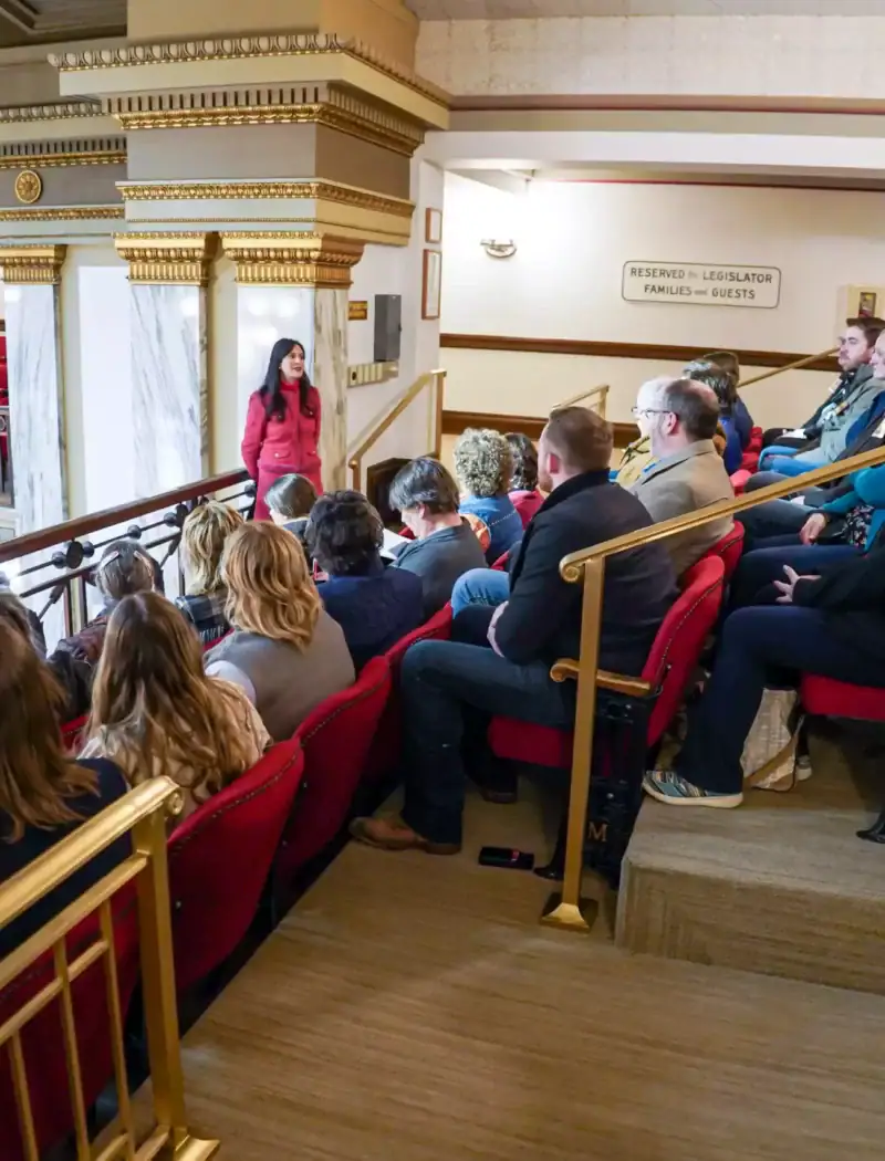 A woman stands in front of a seated audience in a balcony area of an ornate building, possibly a theater or government hall, speaking to the group. The audience listens attentively in red-cushioned seats.