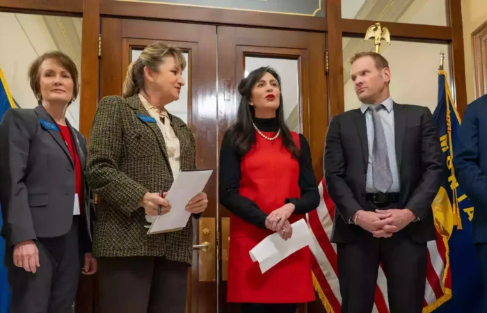 Six professionally dressed people stand in a formal room with wooden doors and flags, engaged in conversation; one woman in a red dress, possibly Secretary Christi Jacobsen, speaks as others—likely discussing U.S. citizens voting or Montana Legislature matters—listen attentively.