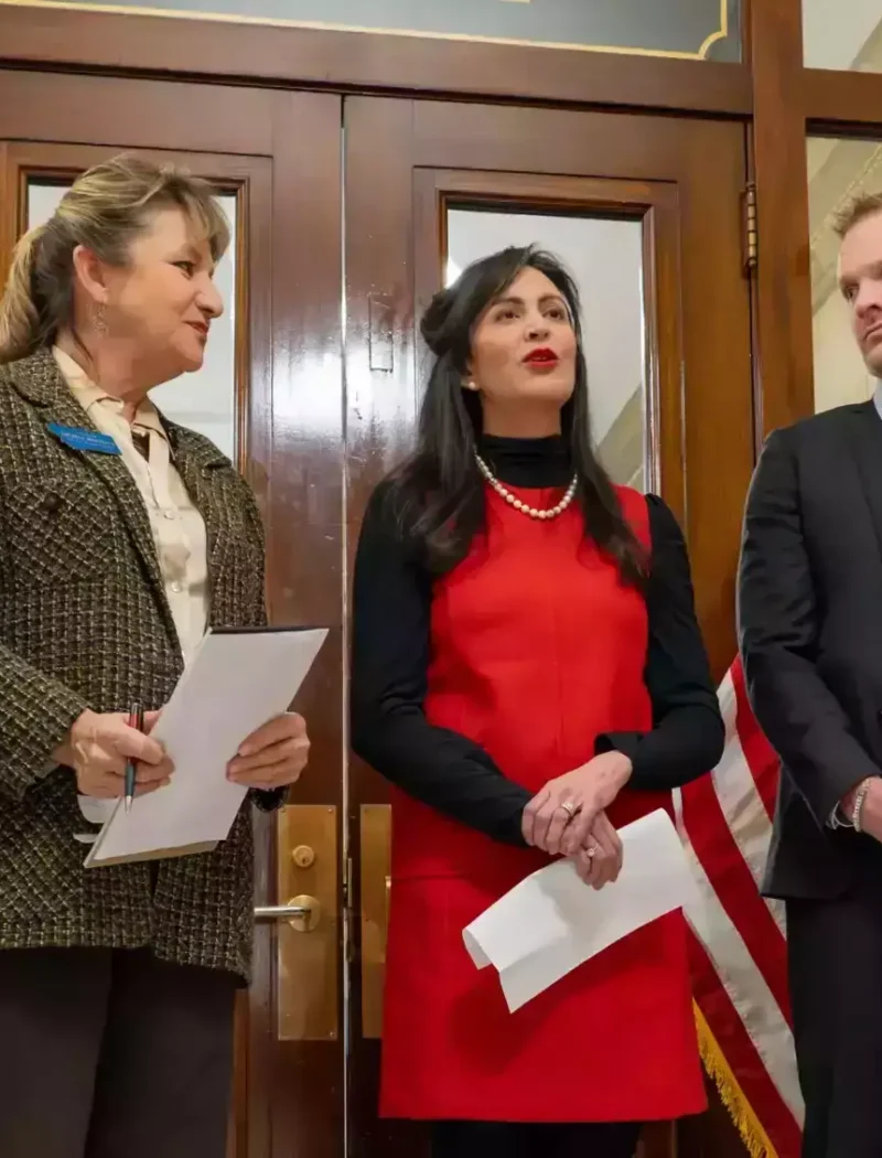 Six professionally dressed people stand in a formal room with wooden doors and flags, engaged in conversation; one woman in a red dress, possibly Secretary Christi Jacobsen, speaks as others—likely discussing U.S. citizens voting or Montana Legislature matters—listen attentively.