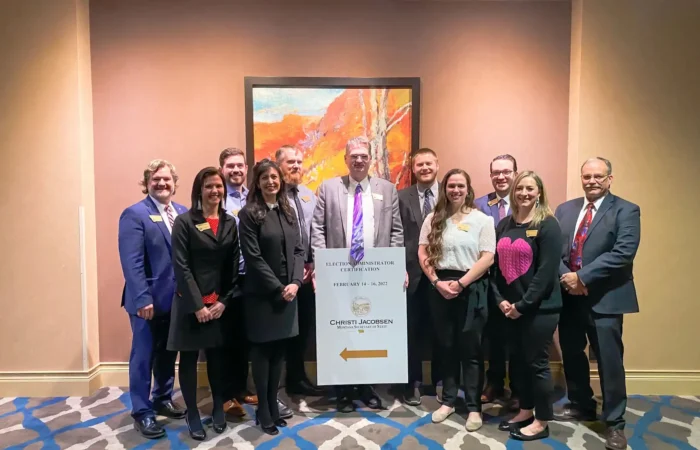 Montana Secretary of State's Office hosts Election Administrator Certification training 6 A group of twelve people, dressed in business attire, stand together in a hotel conference room, smiling for a photo beside a sign directing to an event. A colorful painting hangs on the wall behind them.