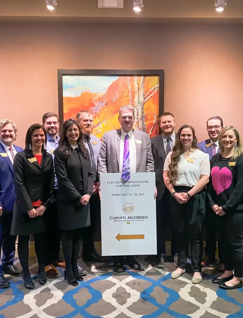 A group of twelve people, dressed in business attire, stand together in a hotel conference room, smiling for a photo beside a sign directing to an event. A colorful painting hangs on the wall behind them.