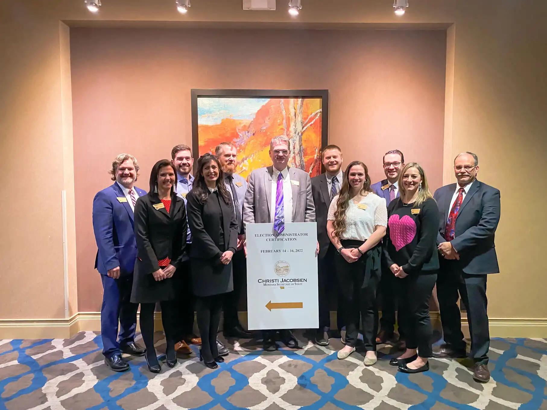 A group of twelve people, dressed in business attire, stand together in a hotel conference room, smiling for a photo beside a sign directing to an event. A colorful painting hangs on the wall behind them.
