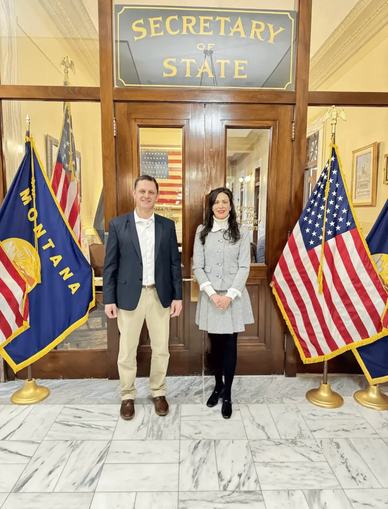 Two people stand side by side in front of the Secretary of State office doors, flanked by Montana state and U.S. flags on each side. The setting appears formal, with marble floors and framed items on the walls.