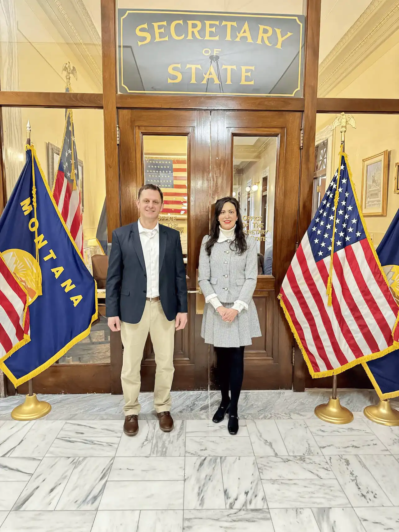 Two people stand side by side in front of the Secretary of State office doors, flanked by Montana state and U.S. flags on each side. The setting appears formal, with marble floors and framed items on the walls.