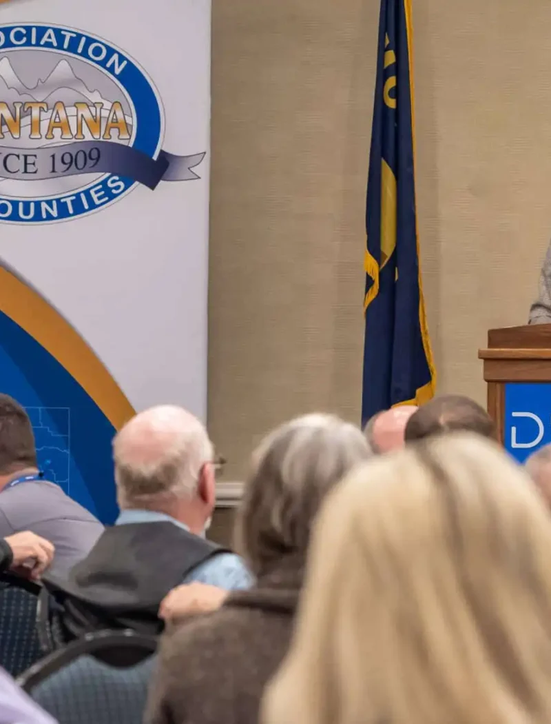A woman, possibly Secretary Christi Jacobsen of the SOS Office, speaks at a podium during an indoor event. Attendees face her as she stands before a Montana Association of Counties sign and a flag, underscoring the official setting.