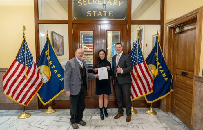 Three people stand smiling in front of the Montana Secretary of State office, holding a resolution. They are flanked by two U.S. flags and two Montana state flags on each side. Secretary Christi Jacobsen stands in the middle, proudly holding the document.