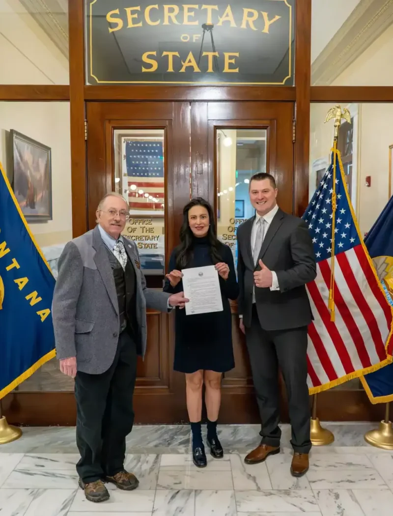 Three people stand smiling in front of the Montana Secretary of State office, holding a resolution. They are flanked by two U.S. flags and two Montana state flags on each side. Secretary Christi Jacobsen stands in the middle, proudly holding the document.