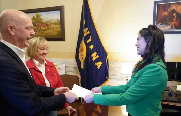 Secretary Jacobsen: 'Candidates for Governor, U.S. Senate and House, highlight week of candidate filing' 3 Three people stand indoors exchanging a document; a man in a suit and a woman in a red jacket face a woman in a green blazer. A Montana state flag and two paintings are visible in the background.