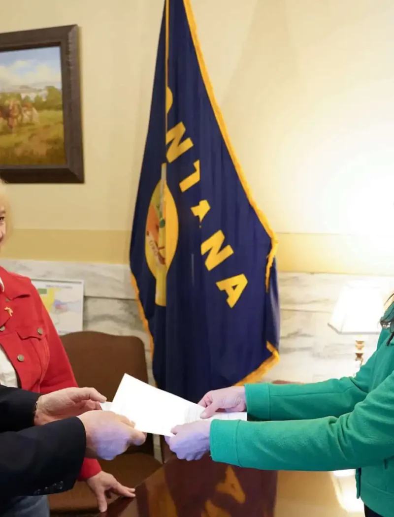 Three people stand indoors exchanging a document; a man in a suit and a woman in a red jacket face a woman in a green blazer. A Montana state flag and two paintings are visible in the background.
