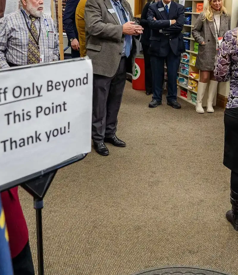 A group of people stand and talk beyond a sign that reads Staff Only Beyond This Point Thank you! An American flag is visible in the foreground, and the setting appears to be indoors, possibly a library or office.
