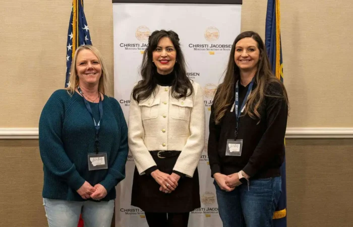 Secretary Christi Jacobsen: 'All 56 Montana counties have completed required third-party security assessments' 7 Three women stand smiling in front of a banner and between two flags. The woman in the center, Christi Jacobsen, wears a white jacket. The background banner features official logos and text related to Montana counties and security assessments.
