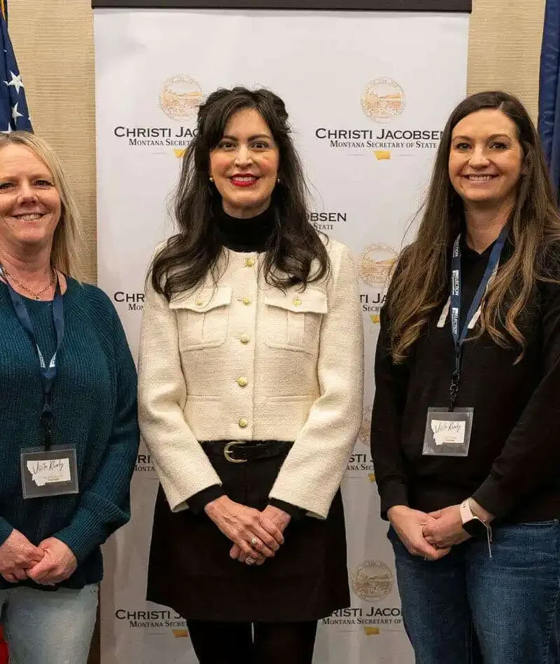 Three women stand smiling in front of a banner and between two flags. The woman in the center, Christi Jacobsen, wears a white jacket. The background banner features official logos and text related to Montana counties and security assessments.